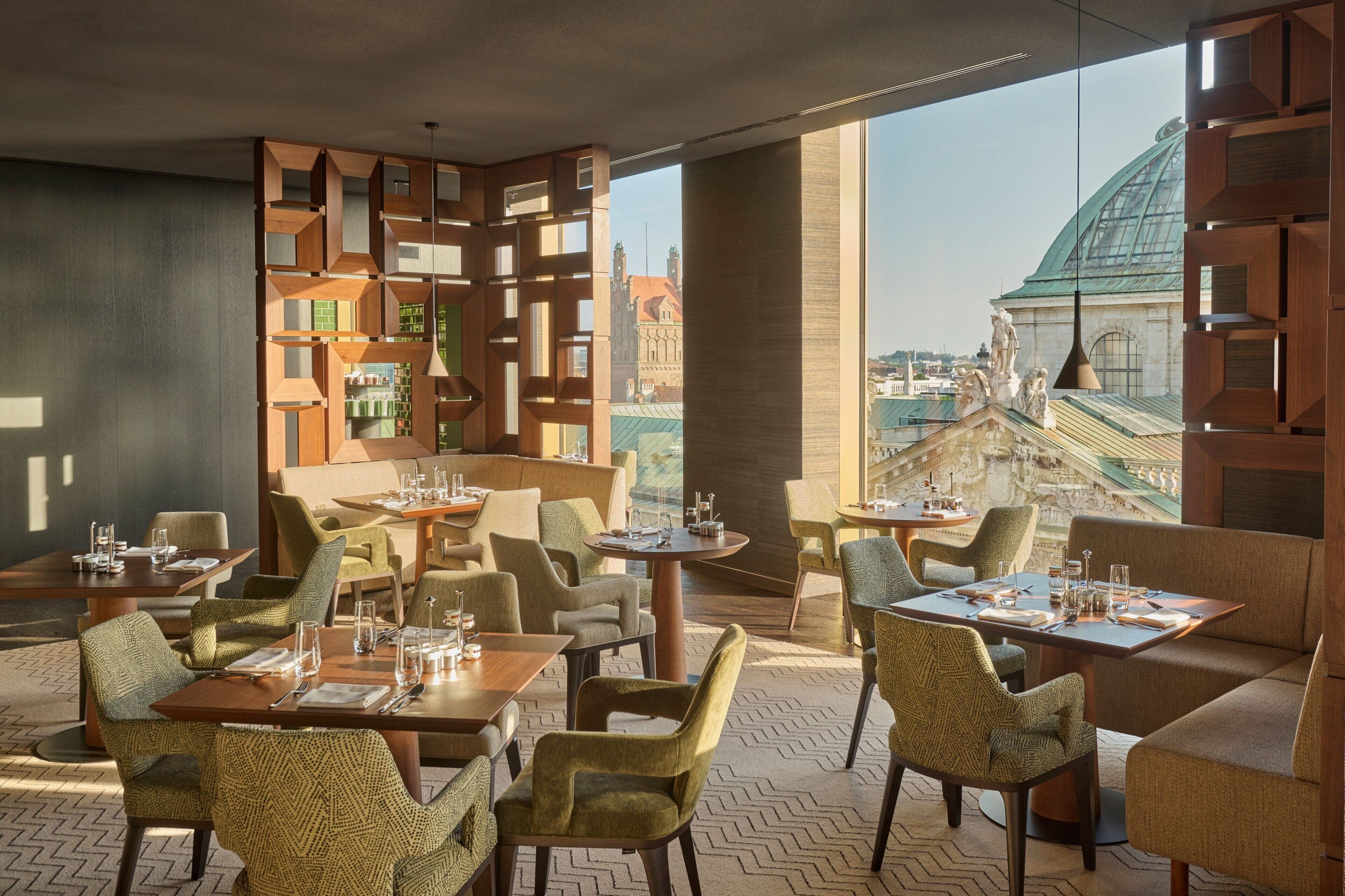 A modern restaurant with a view of historic buildings, featuring geometric wooden partitions, comfortable seating, and a herringbone-patterned floor.