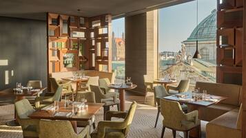 A modern restaurant with a view of historic buildings, featuring geometric wooden partitions, comfortable seating, and a herringbone-patterned floor.