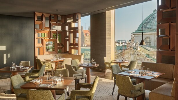 A modern restaurant with a view of historic buildings, featuring geometric wooden partitions, comfortable seating, and a herringbone-patterned floor.