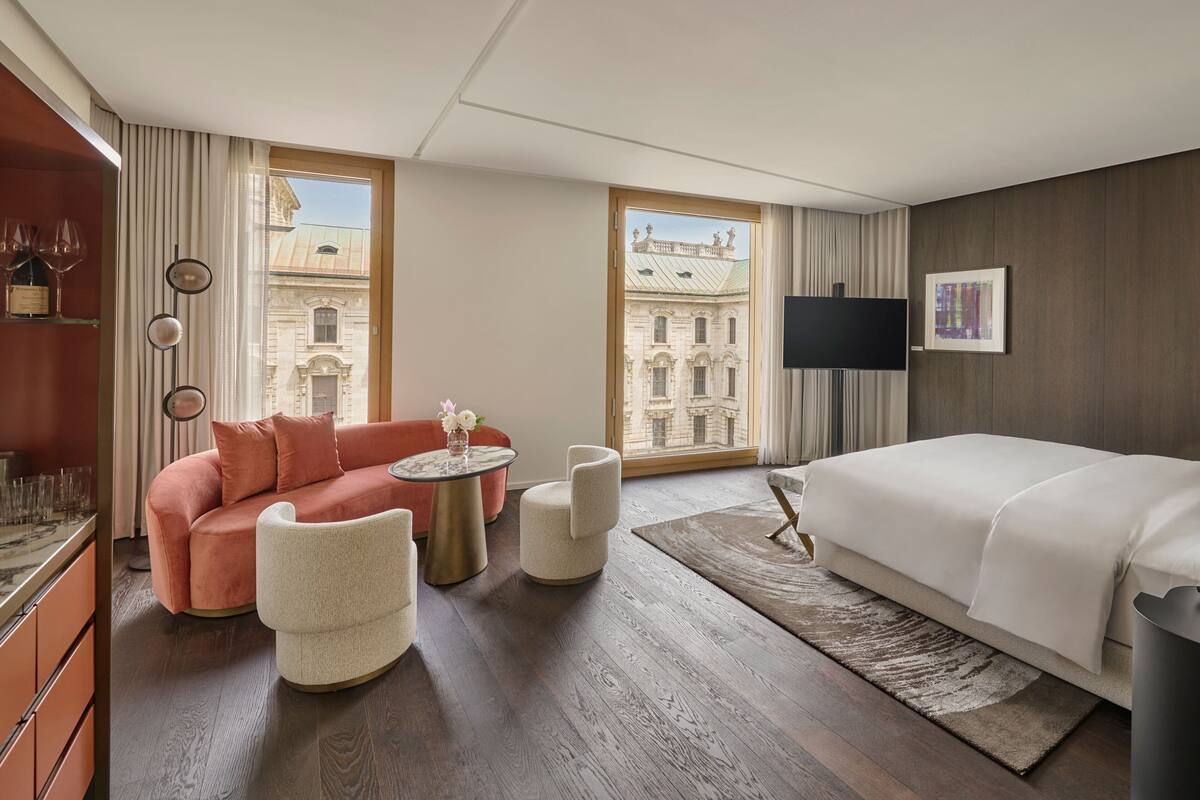 A hotel room with a bed, a sofa, a chair, a small table, and a TV. There are curtains and a window with a view of a building.