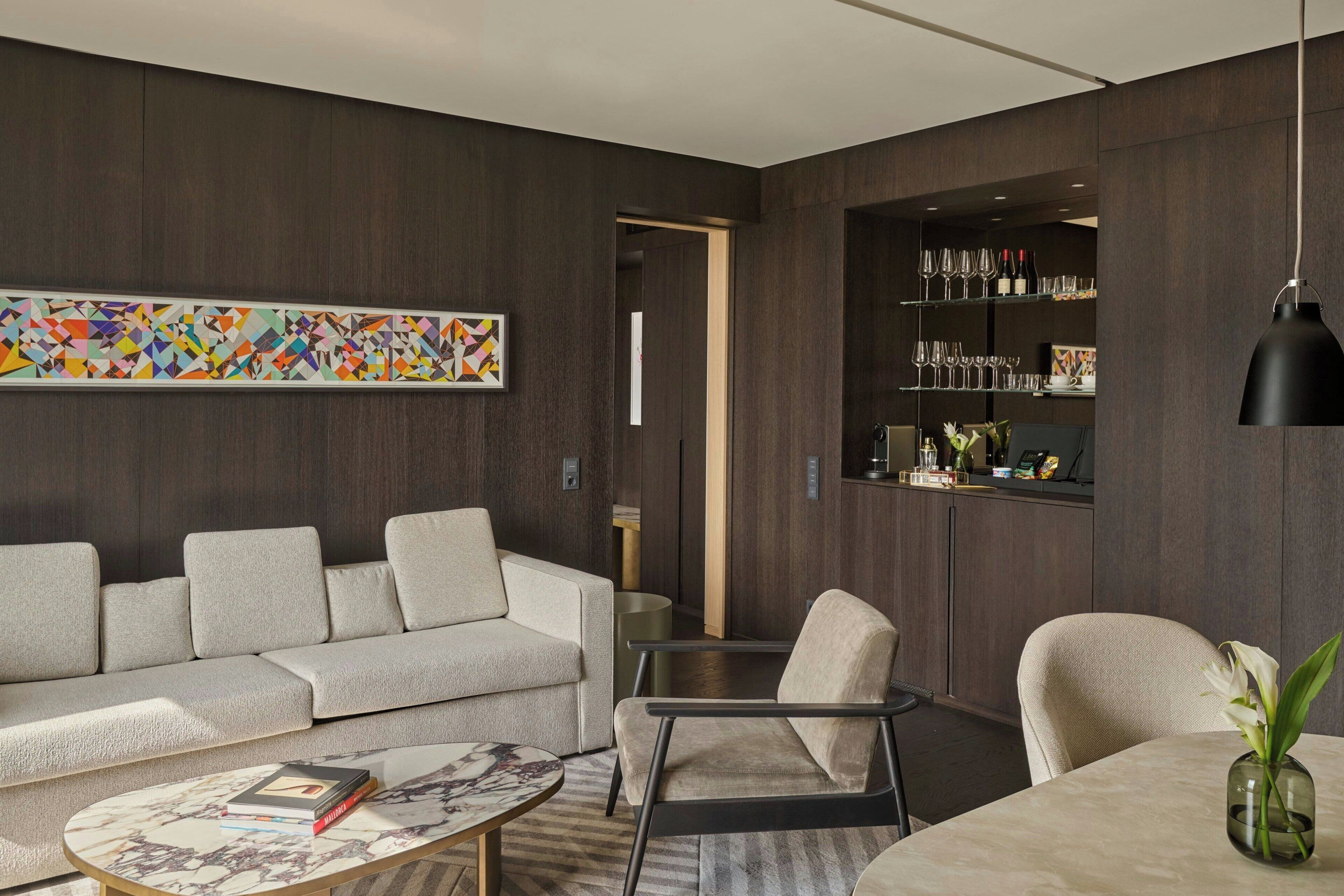 Una sala moderna con sofá, sillones, una mesa de centro y una mesa de comedor. Se ve una estantería con copas de vino y un cuadro en la pared.