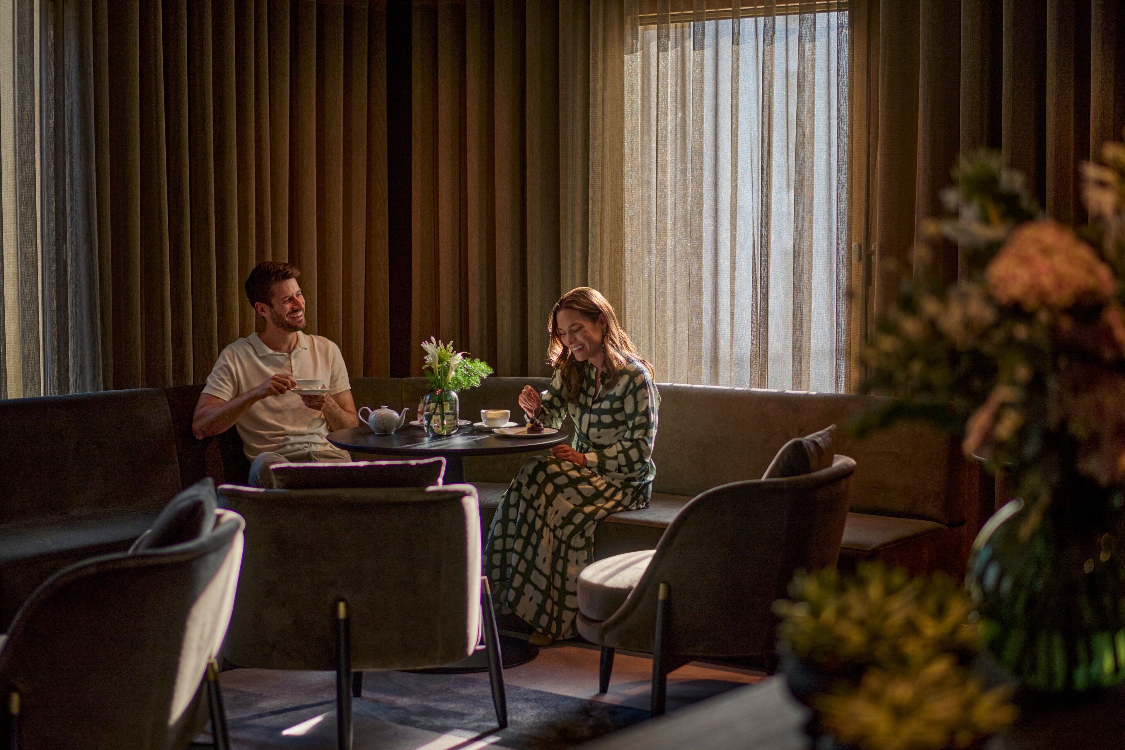 A couple seated at a table in a modern lounge area with a sofa, chairs, and a vase of flowers.