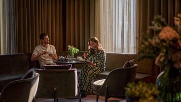 A couple seated at a table in a modern lounge area with a sofa, chairs, and a vase of flowers.
