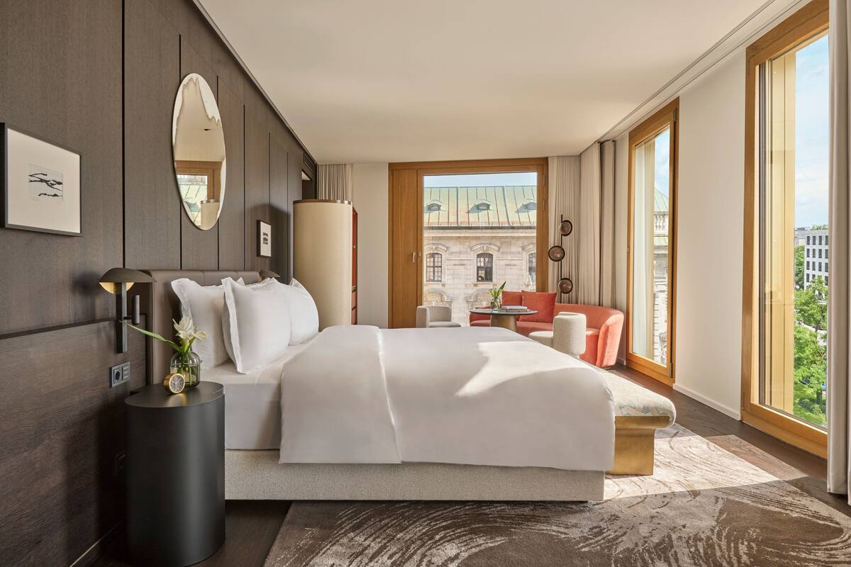 A modern hotel room with a large bed, a sitting area with a red sofa, a round table, and a view of a historic building through large windows.