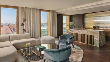 Una habitación de hotel moderna con sofá, sillas, una pequeña mesa, zona de cocina y ventanas amplias con vista a la ciudad.