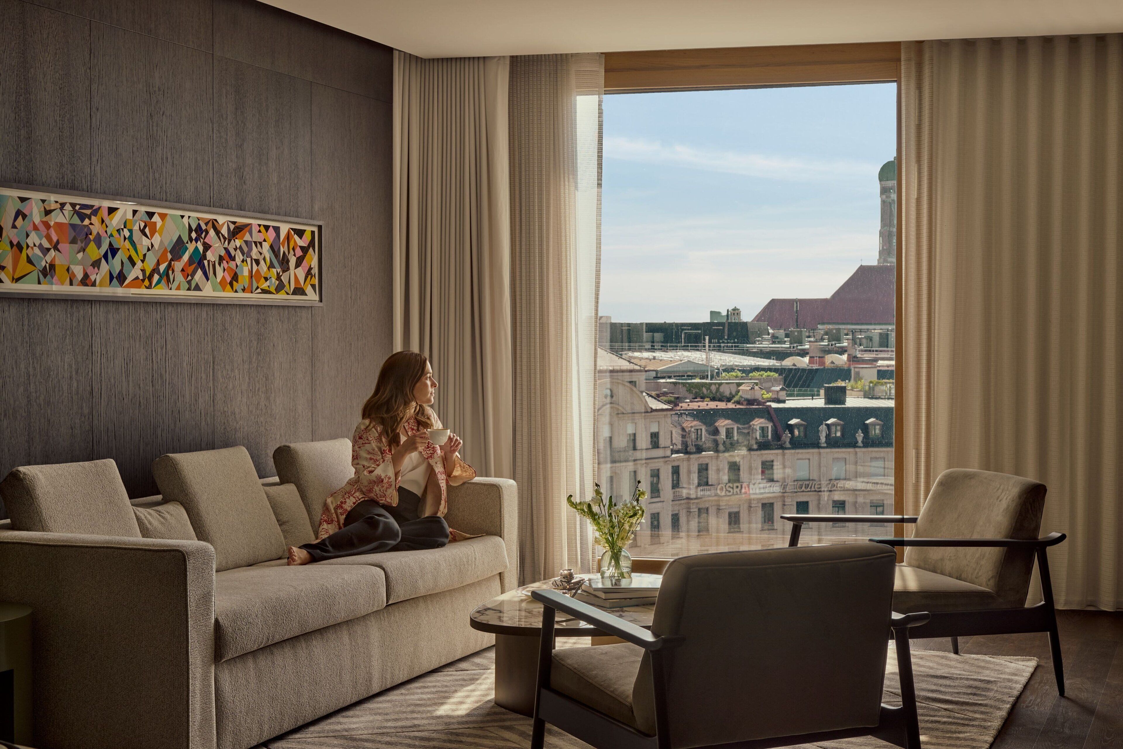 Una sala de estar moderna con sofá, sillones y una mesa de centro. Un ventanal ofrece vistas a edificios y una torre.