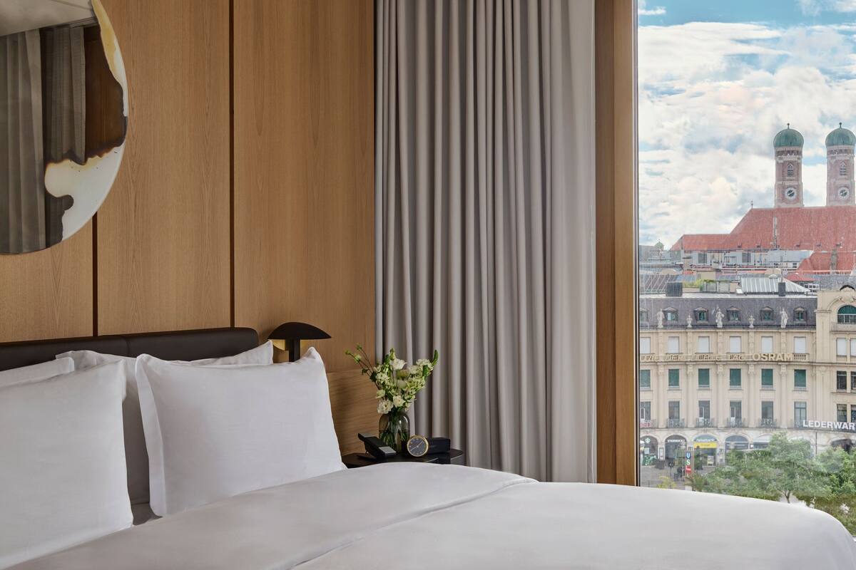 A hotel room with a large bed, a wooden headboard, a nightstand with a clock and a vase of flowers, and a view of a cityscape with buildings and a clock tower.