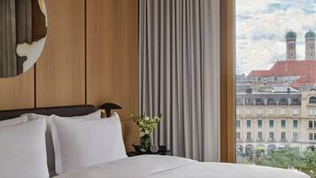 Una habitación de hotel con una cama grande, un cabecero de madera, una mesita de noche con un reloj y un florero, y vistas a un paisaje urbano con edificios y una torre del reloj.