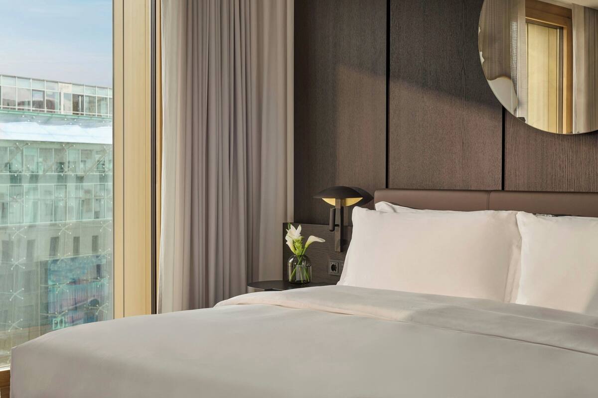 A hotel room with a large bed, a nightstand with a lamp, a vase with flowers, and a view of a cityscape through the window.