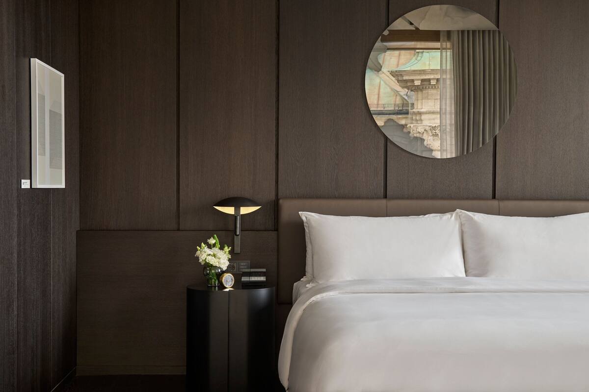 A modern hotel room with a large bed, a round mirror, a nightstand with a lamp, and a view of the city through the window.