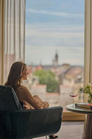 A person sitting in a chair by a window with a view of buildings and a table with a book and a glass.