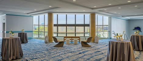 A spacious room with a patterned carpet, round tables with draped covers, and a large window offering a view of the cityscape.
