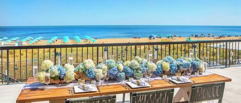 A wooden table set with glassware and flowers, overlooking a beach with umbrellas.