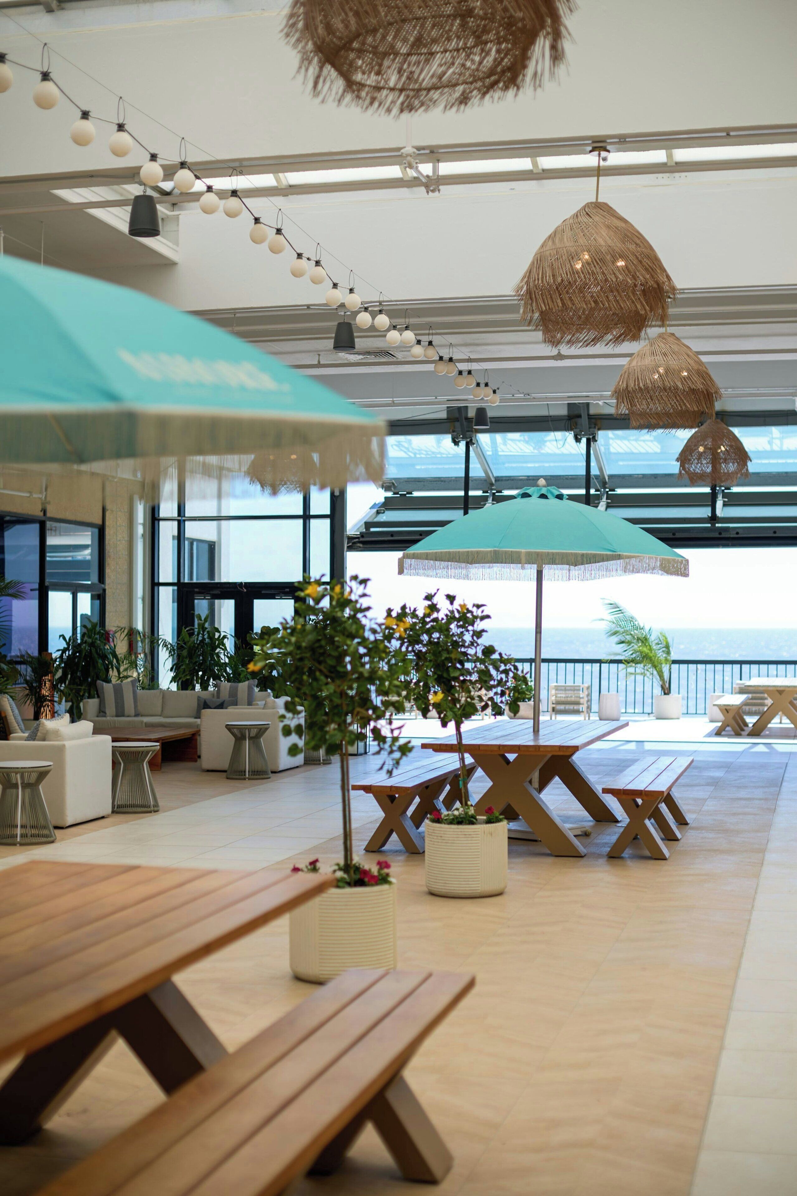 Un espace intérieur spacieux, doté de bancs, de tables et de parasols en bois, de grandes fenêtres et offrant une vue sur la mer.