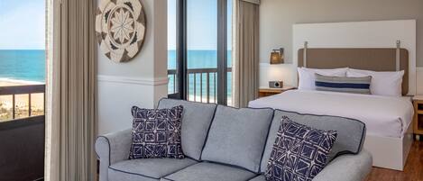 A hotel room with a balcony, a grey sofa with patterned cushions, a bed with white linens, and a wall-mounted decorative plate.
