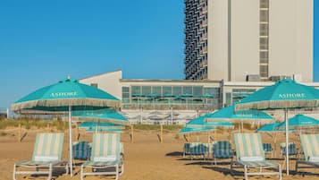 Sillas de playa y sombrillas con la marca 'ASHORE', un edificio moderno con fachada estampada y un cielo azul despejado.