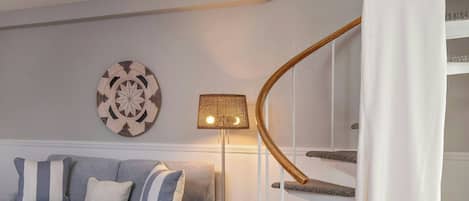 Un salón moderno con un sofá gris, una mesa de centro de madera, un vestido blanco colgado, una escalera de caracol y un plato decorativo en la pared.