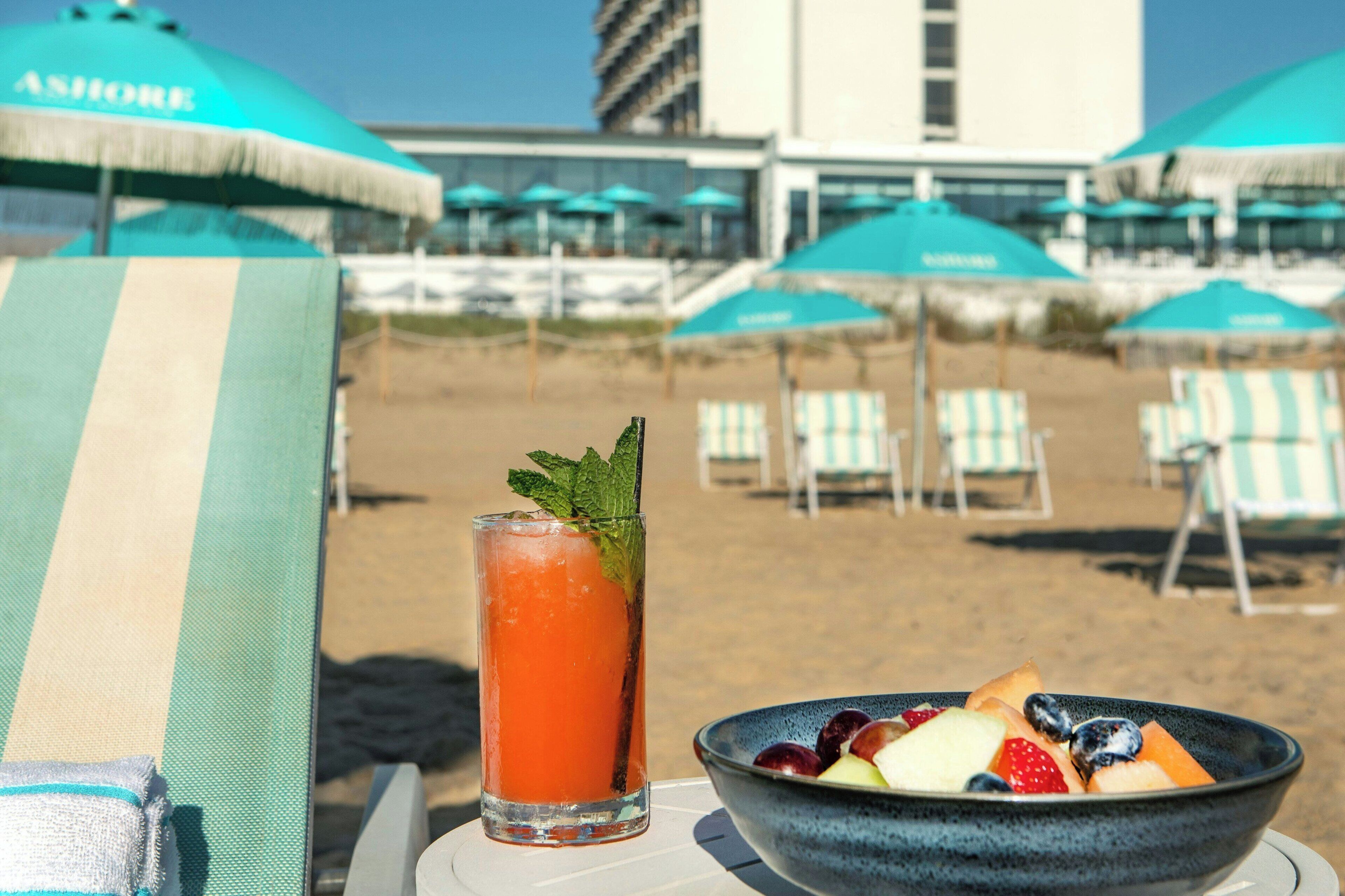 Una spiaggia con un cocktail, una ciotola di frutta e sedie a sdraio a righe sotto gli ombrelloni.