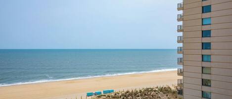 A beachfront hotel with a view of the ocean, a sandy beach, and a parking area with several air conditioning units.