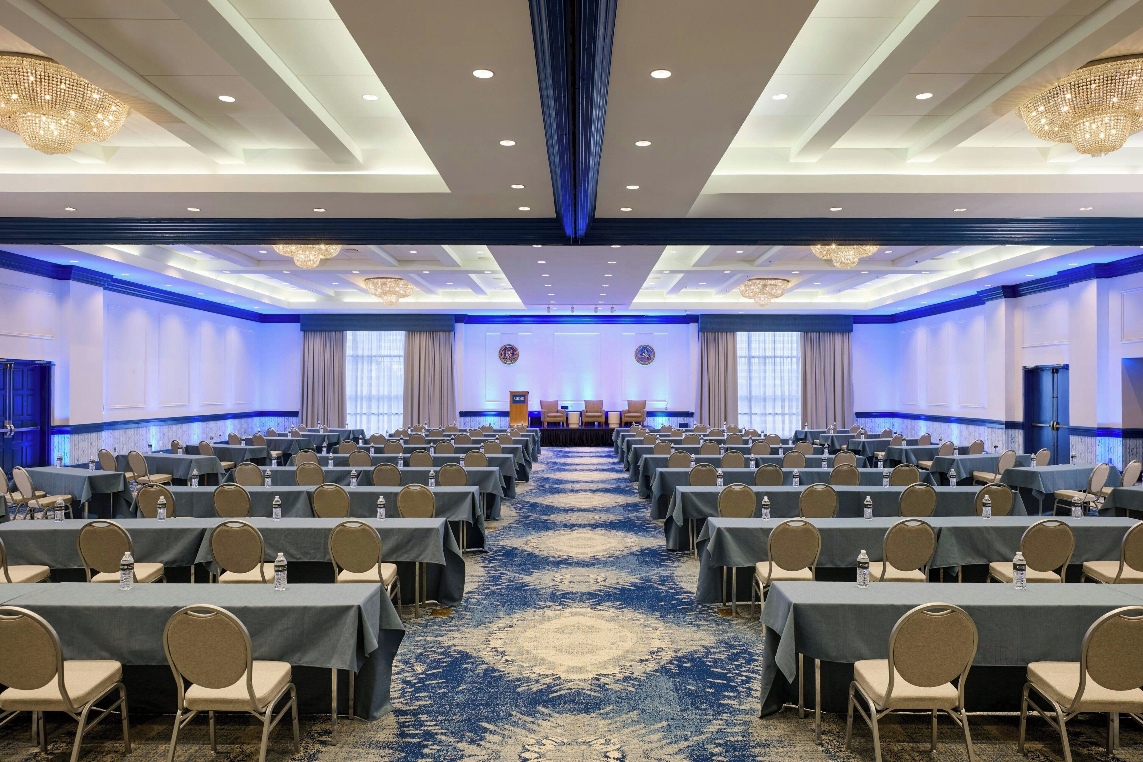 A conference room with rows of tables and chairs, a stage with podiums, and chandeliers.