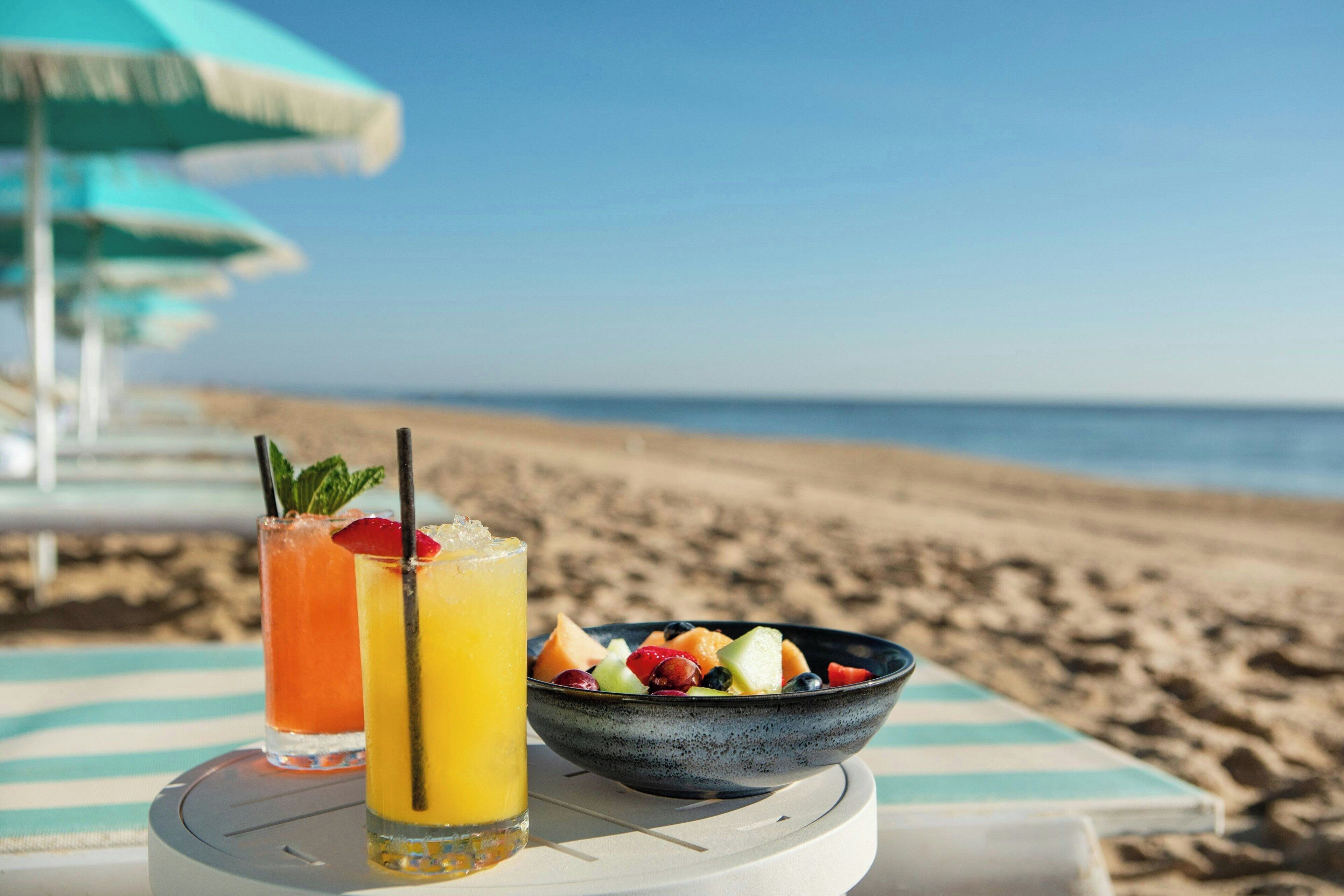 Mesa à beira-mar com uma cesta de frutas e duas bebidas, um guarda-sol listrado e vista desobstruída para o oceano.