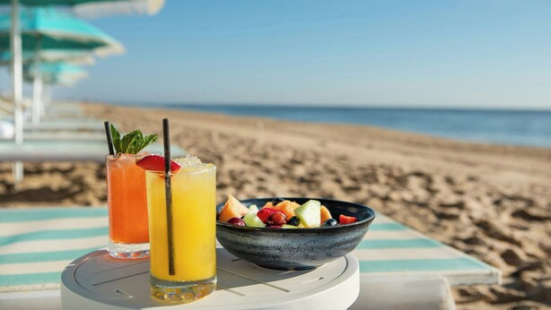 Een strandtafel met een fruitschaal en twee drankjes, een gestreepte parasol en een helder uitzicht op de oceaan.
