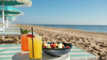 A beachside table with a fruit bowl and two drinks, a striped umbrella, and a clear view of the ocean.
