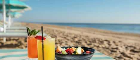 A beachside table with a fruit bowl and two drinks, a striped umbrella, and a clear view of the ocean.