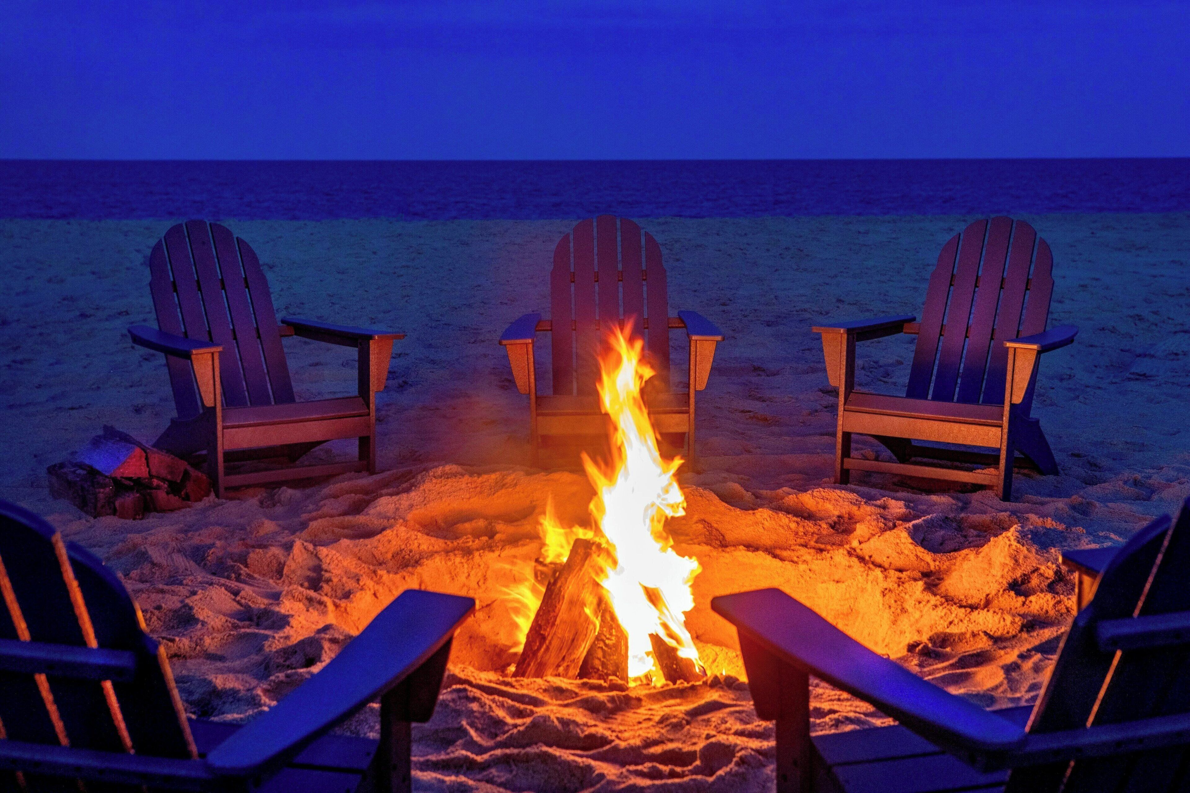 Escena de playa con tres sillas Adirondack dispuestas alrededor de una fogata.