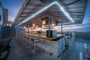 Un área de bar al aire libre con un espacio de asientos cubierto, una barra y vista a la playa al atardecer.