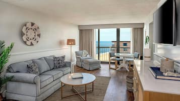 A modern living room with a sofa, coffee table, and a TV. There is a balcony with a view of the sea.