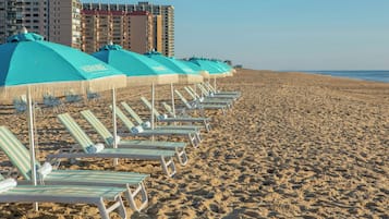 Una playa con filas de tumbonas y sombrillas, una orilla arenosa y edificios altos al fondo.