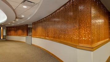 A spacious hallway with a curved wooden wall featuring intricate perforations, a carpeted floor, and a recessed ceiling with lighting.