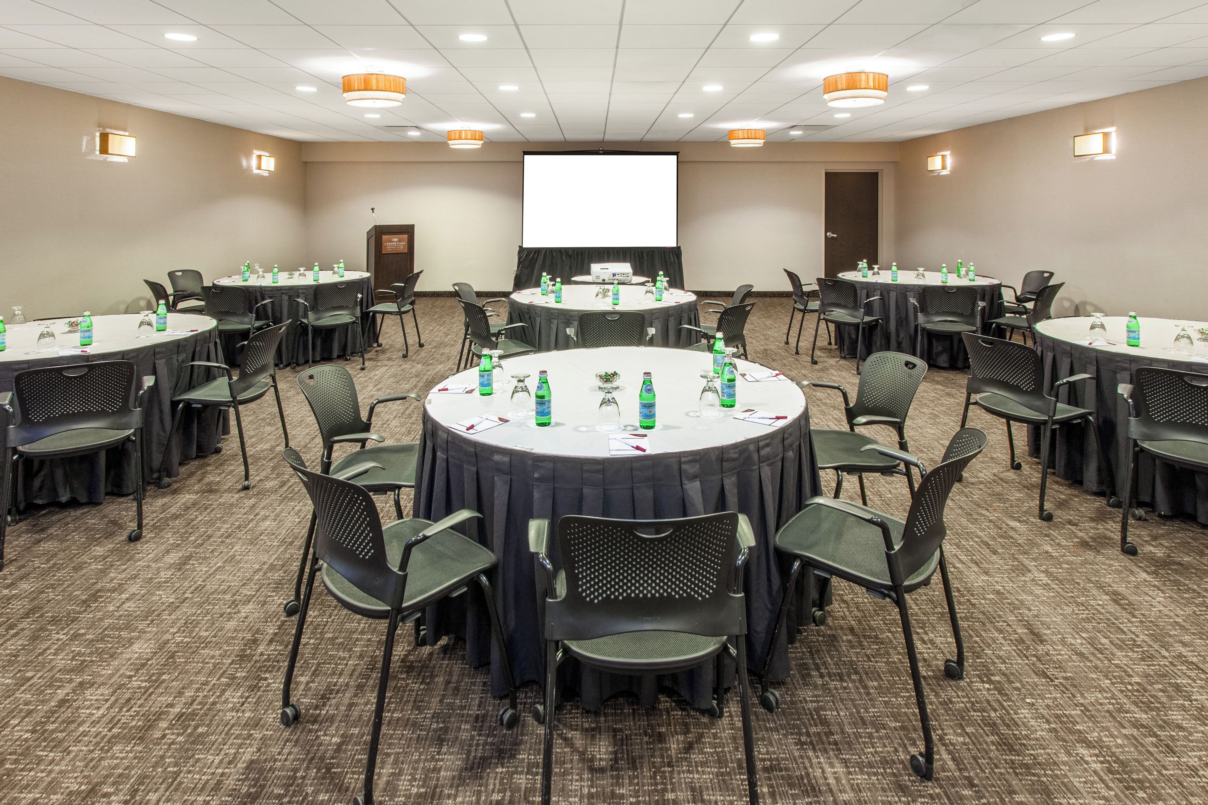 Une salle de conférence dotée de plusieurs tables rondes, chacune étant entourée de chaises, et équipée de bouteilles d’eau et de verres.