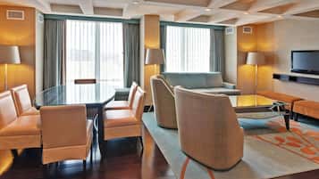 A hotel room with a dining table, chairs, a sofa, a TV, and a large window with curtains.