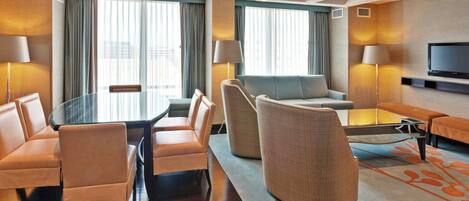 A hotel room with a dining table, chairs, a sofa, a TV, and a large window with curtains.