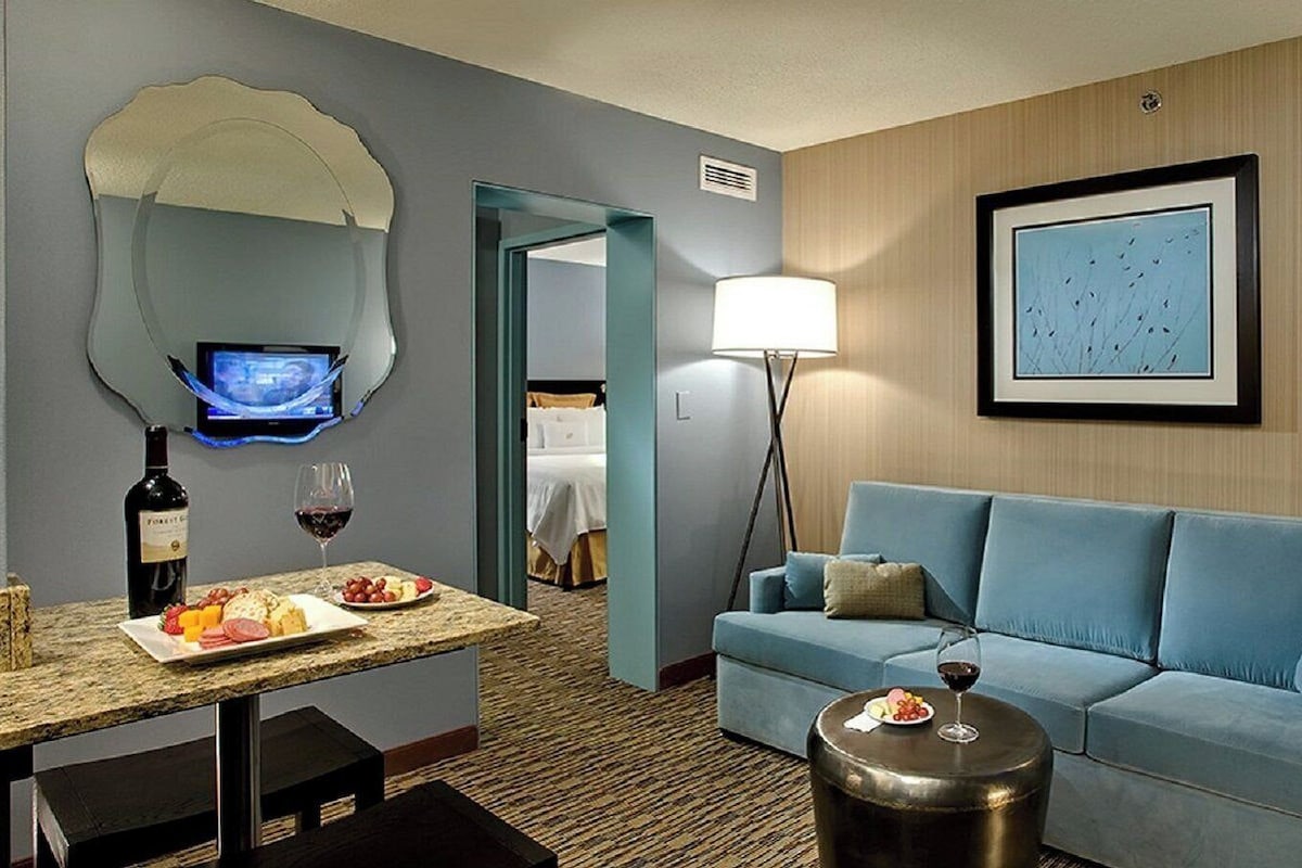 A hotel room with a blue sofa, a small round table, a TV on a wall, a dining table with a bottle of wine and a glass, and a framed picture on the wall.