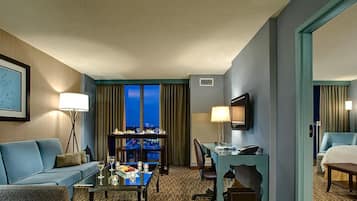 A hotel room with a blue sofa, a dining table with a view, a desk with a chair, and a bed.