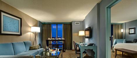 A hotel room with a blue sofa, a dining table with a view, a desk with a chair, and a bed.