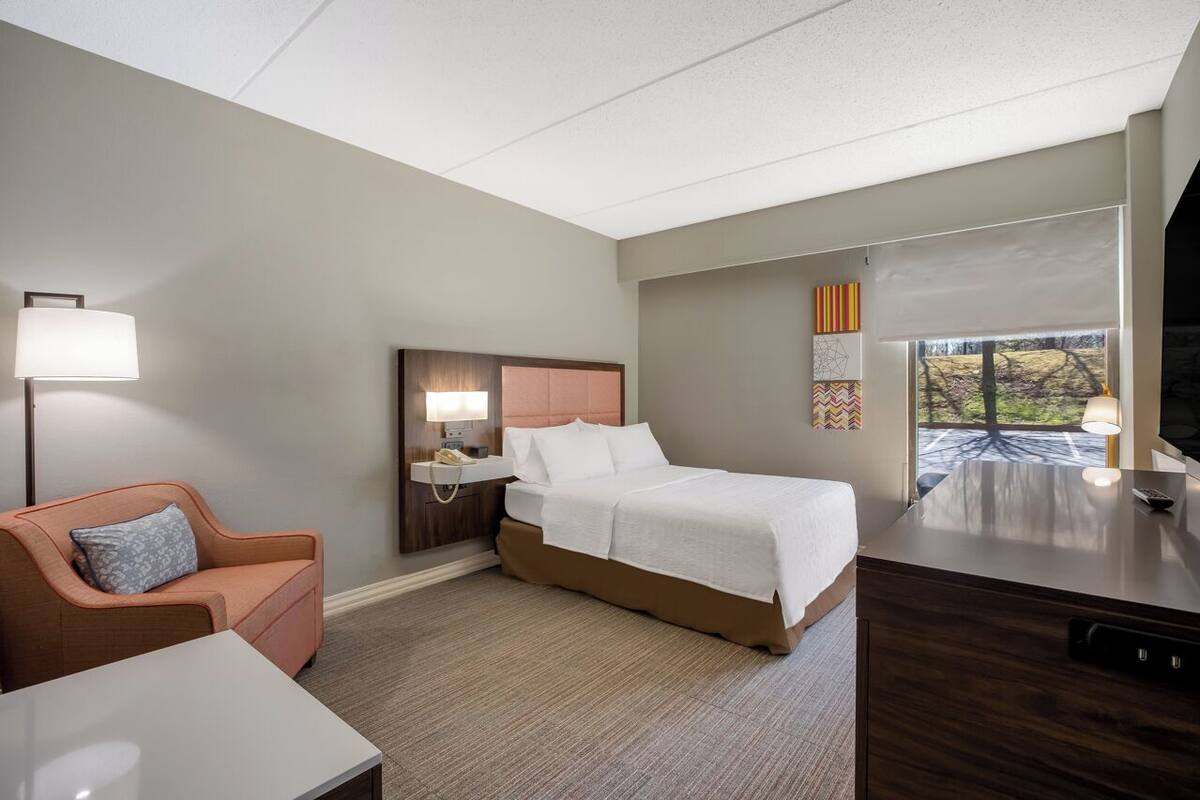 A hotel room with a bed, a desk, a chair, a nightstand, and a window with a view of trees.