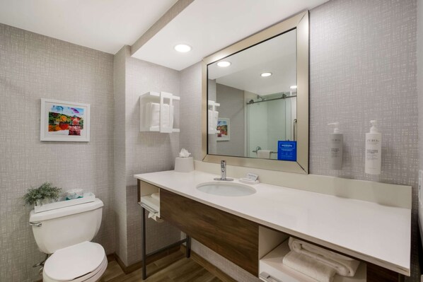 A modern bathroom with a white sink, large mirror, and a toilet. There is a framed picture on the wall and a small plant on the toilet.