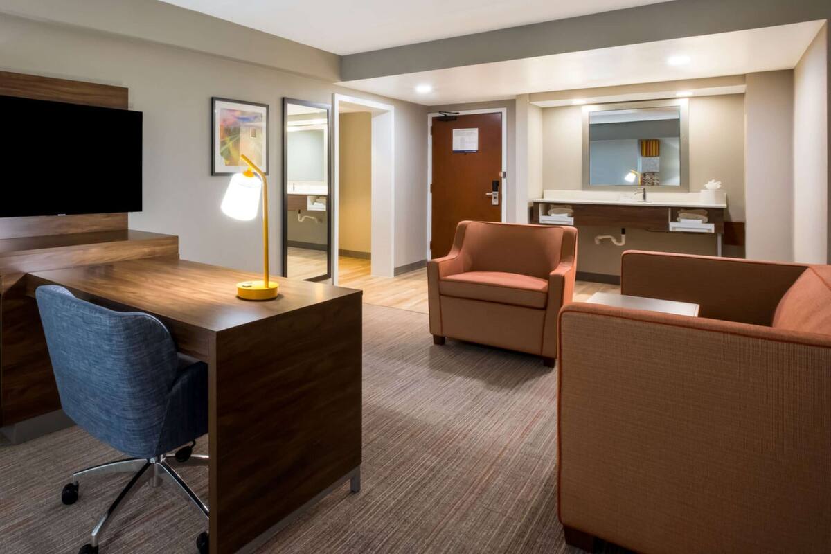 A hotel room with a wooden desk, a blue office chair, two orange armchairs, a small table with a lamp, a large mirror, and a door leading to a bathroom.