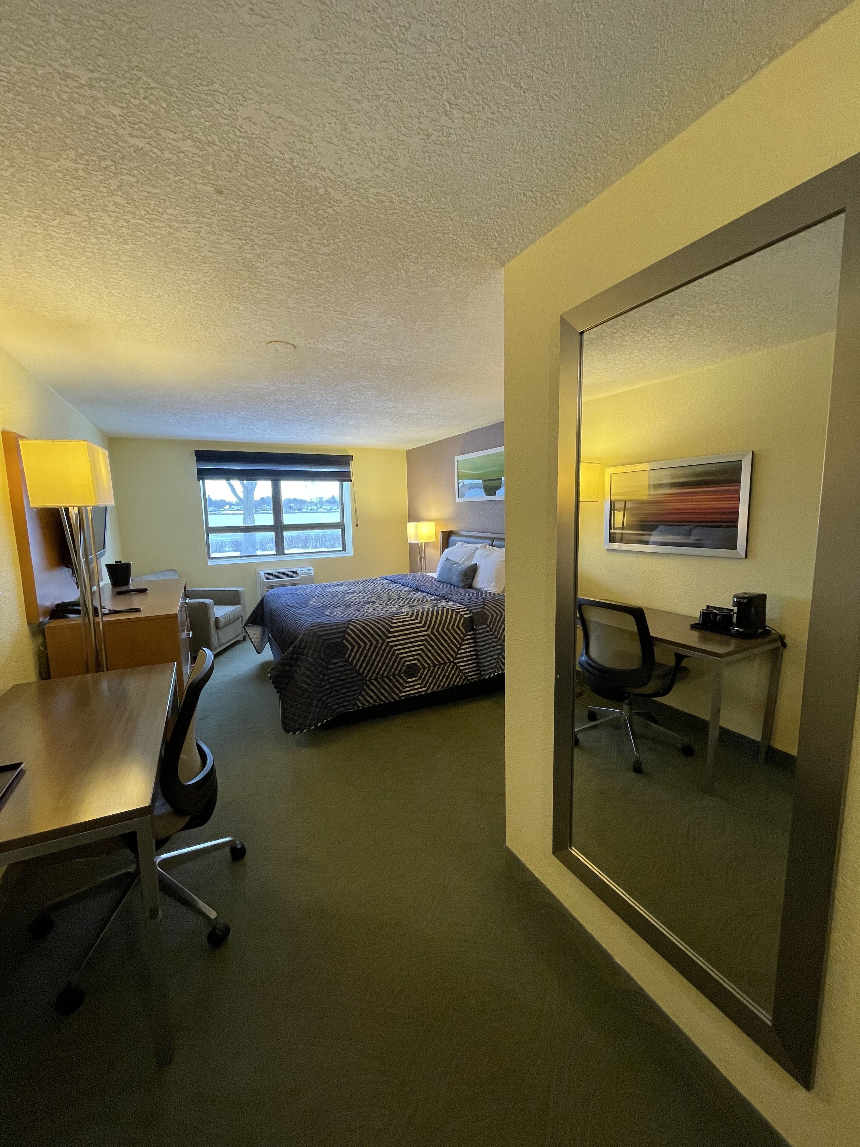 Une chambre d’hôtel comprenant un lit, un bureau, une chaise, une lampe, une fenêtre et un miroir.