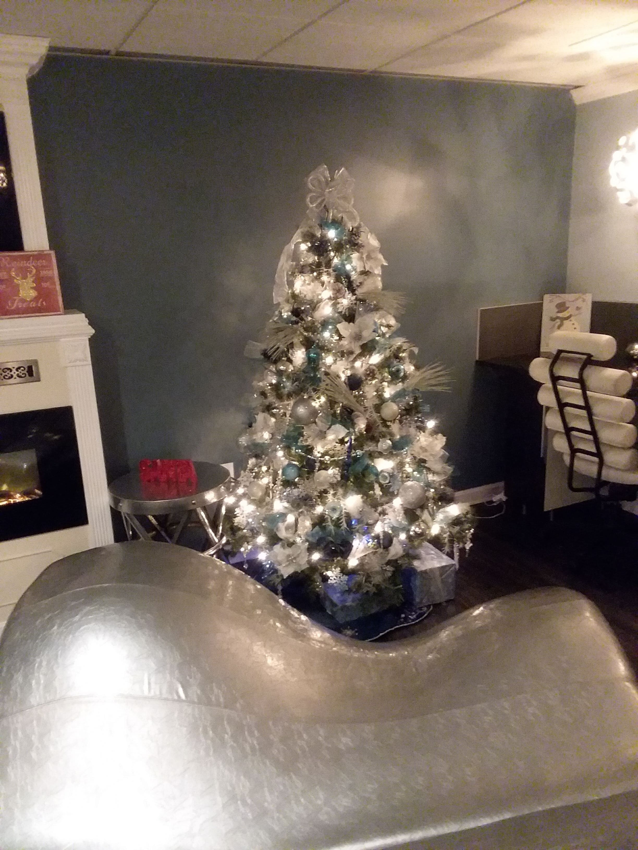 A Christmas tree in a room with a dark blue wall, a white fireplace, and a dining area with a chair and table.