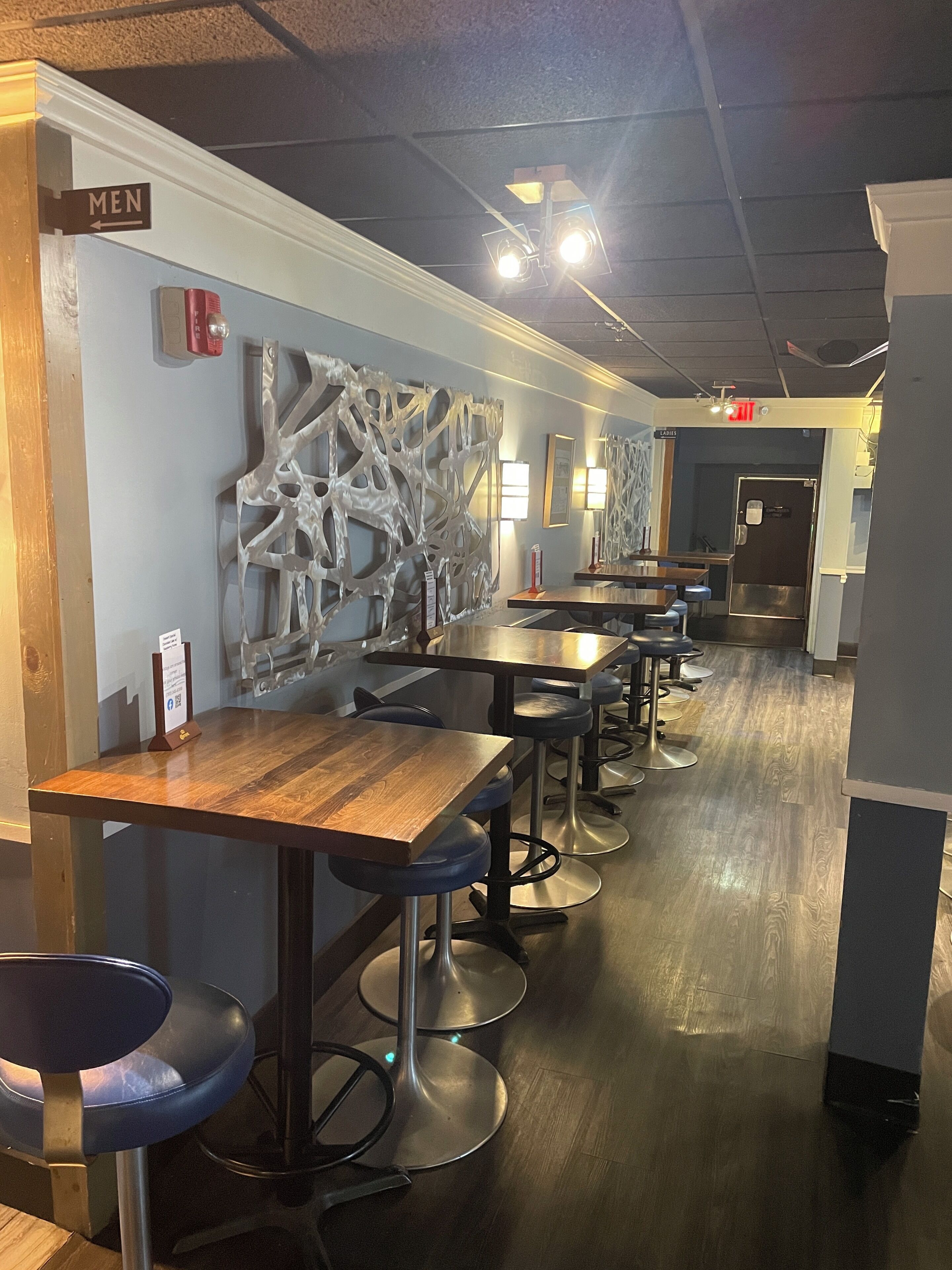 Interior moderno de restaurante con mesas de madera, taburetes de bar azules, una pared decorativa y señales direccionales para hombres y mujeres.