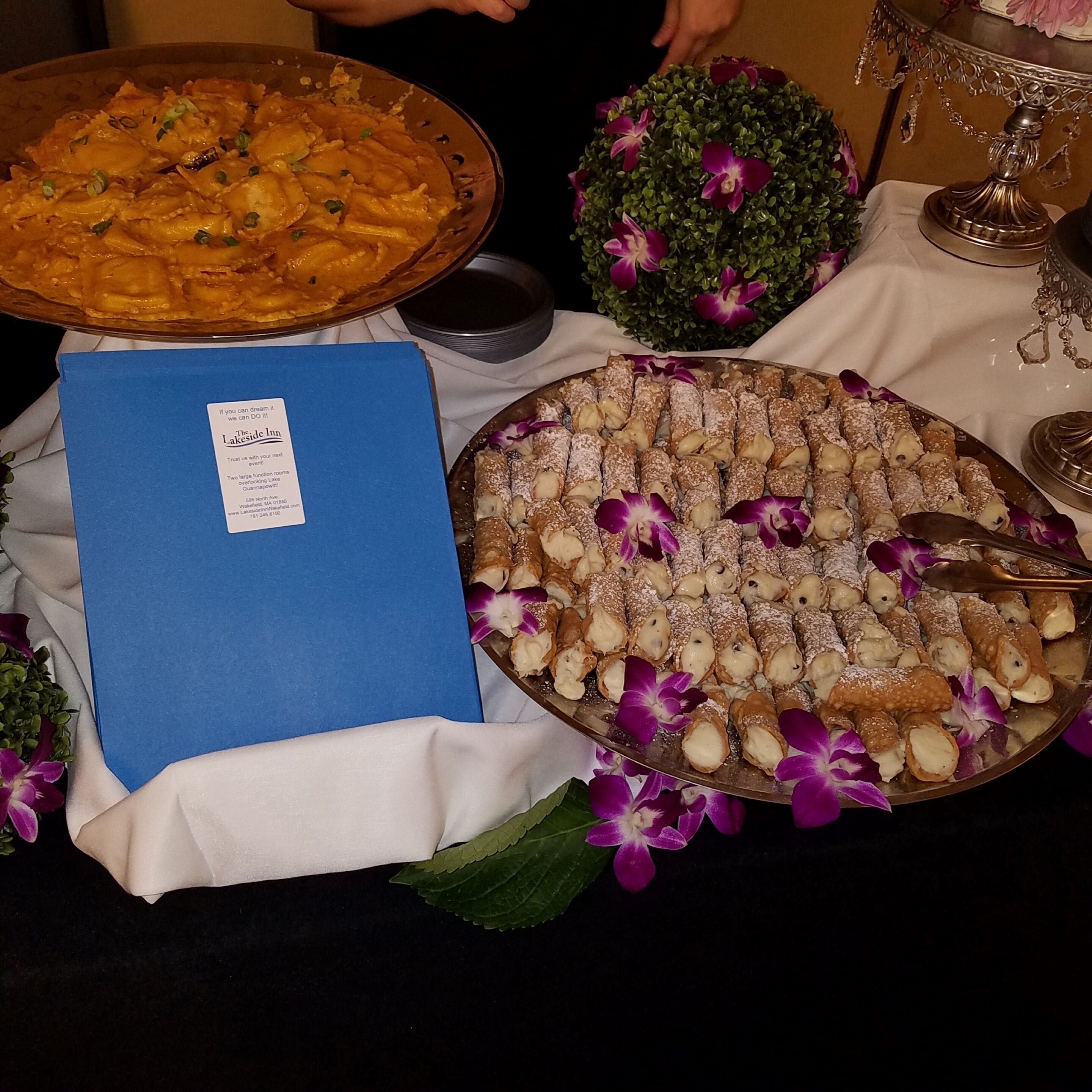 Uma mesa com uma grande bandeja de cannoli, uma pasta azul e um arranjo decorativo de flores roxas.