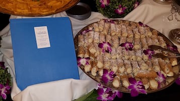 Uma mesa com uma grande bandeja de cannoli, uma pasta azul e um arranjo decorativo de flores roxas.