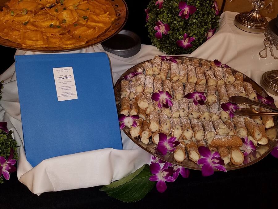 Ein Tisch mit einer großen Platte mit Cannoli, einer blauen Ordner und einer dekorativen Anordnung aus violetten Blumen.
