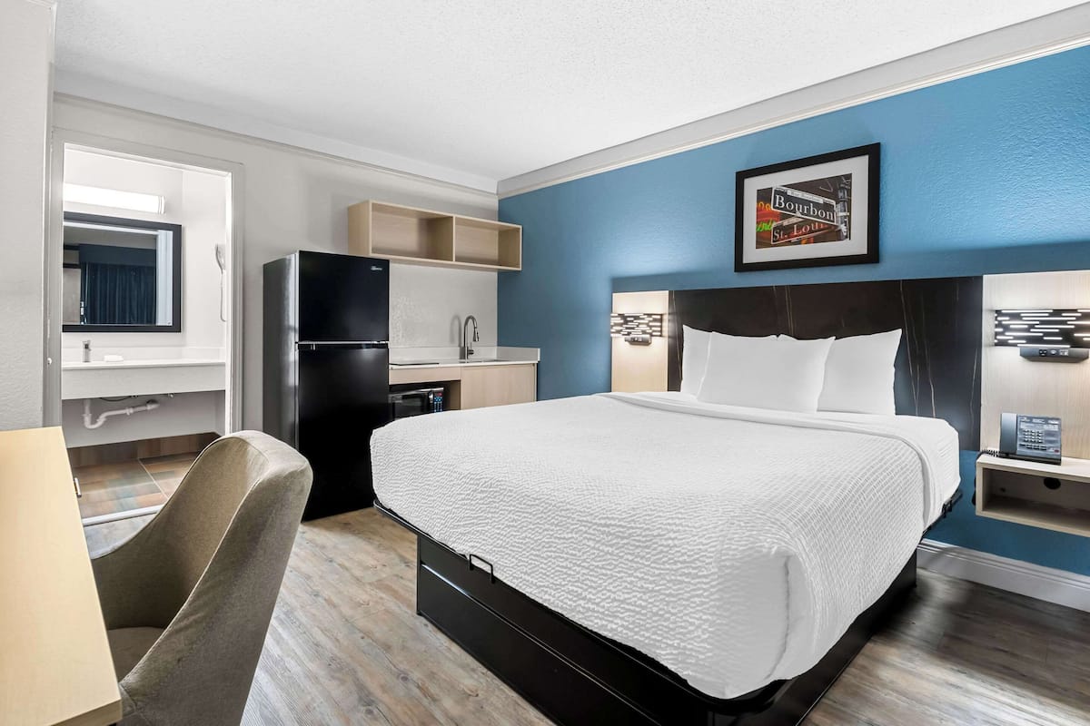 A hotel room with a bed, a desk, a chair, a refrigerator, a microwave, a sink, and a framed picture on the wall.