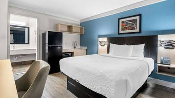 A hotel room with a bed, a desk, a chair, a refrigerator, a microwave, a sink, and a framed picture on the wall.